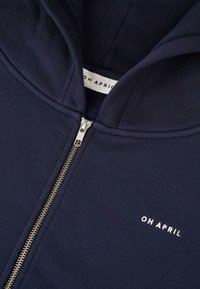 Felpa con zip di colore blu navy realizzata in cotone, caratterizzata da una consistenza morbida, una zip dorata e un'etichetta stampata con la scritta "OH APRIL" in bianco.