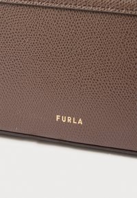 Brūna ādas maks ar teksturētu virsmu. Apakšējā labajā stūrī iegrebts zeltīts "FURLA" logotips. Tīras līnijas un minimālistisks dizains.