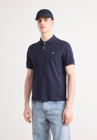 Marinblå kortärmad pikétröja med tvåknappsstängning och liten logotyp på bröstet; kombinerad med ljusblå jeans och en matchande keps.