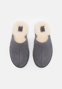 Szürke velúr papucsok tan báránybőr béléssel, slip-on kialakítás, diszkrét varrási részletek és UGG logó a külső oldalán. Lapos gumitalp.