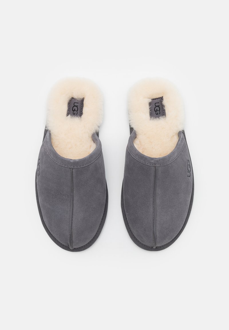 Pantofole in velluto grigio con fodera in pelliccia di pecora tan, design slip-on, dettagli di cucitura sottili e logo UGG sul lato esterno. Suola piatta in gomma.