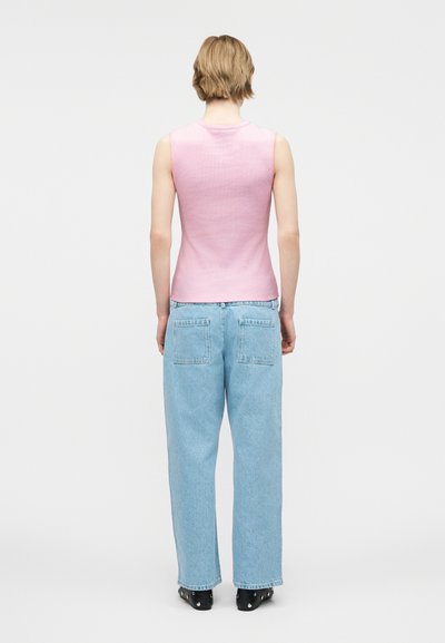 Άτομο με κοντά ξανθά μαλλιά που φοράει αμάνικο ροζ τοπ, ανοιχτό μπλε τζιν και μαύρα παπούτσια, στεκόμενο με την πλάτη προς τα μπρος σε ένα απλό λευκό φόντο.