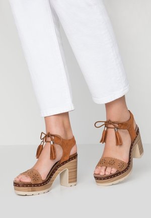 Pieds portant des sandales à talons hauts en daim beige avec liens à la cheville et pompons, associées à un pantalon blanc raccourci, debout sur un sol gris.
