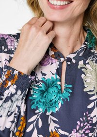 Blusa floreale con scollatura a chiave, polsini elasticizzati e una combinazione di colori vivaci come il teal, il viola e l'arancione su uno sfondo scuro.