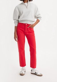 Jeans de mezclilla rojos con corte recto, cinco bolsillos y cierre de botones, combinados con una sudadera gris con cremallera y zapatillas blancas.