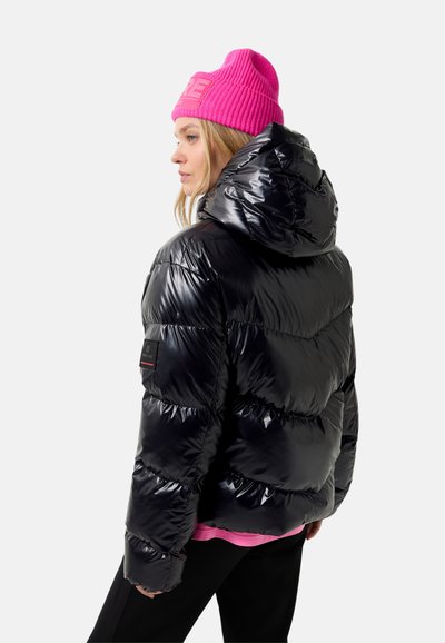 Kvinde i en skinnende sort pufferjakke med hætte, iført en lys pink strikhue og synlig pink skjortefor, vendt mod venstre på en hvid baggrund.
