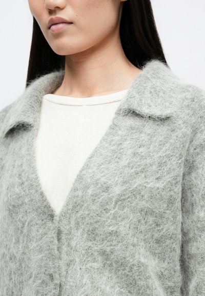 Grijze fleeceachtige cardigan met een kraag met inkeping, gestructureerde stof en een V-hals, gedragen over een licht crèmekleurig topje. Close-up detail van het bovenstuk.