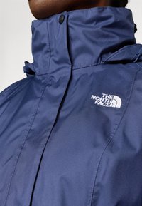 Tamsiai mėlyna vandeniui atspari striukė su aukštu apykakle, kuri turi juodą spaudimo užsegimą ir baltą "The North Face" logotipą kairiajame krūtinės šone.