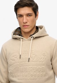 Beige hoodie met reliëflogo, met trekkoord en grijze koordpunten. Gelaagd met een donkergroen geruit overhemd met zichtbare kraag eronder.
