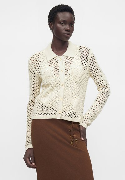 Γυναίκα που φοράει ένα κρεμ πλεκτό ζακέτα με κουμπιά και λουλουδάτα σχέδια και μια καφέ φούστα με δαντέλα στα πλάγια, ποζάρει με το ένα χέρι στη μέση.