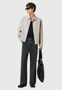 Lichtbeige jas, zwarte t-shirt, donkergrijze wijde jeans, zwarte schoenen en zonnebril. Zwarte tas in de linkerhand vastgehouden.