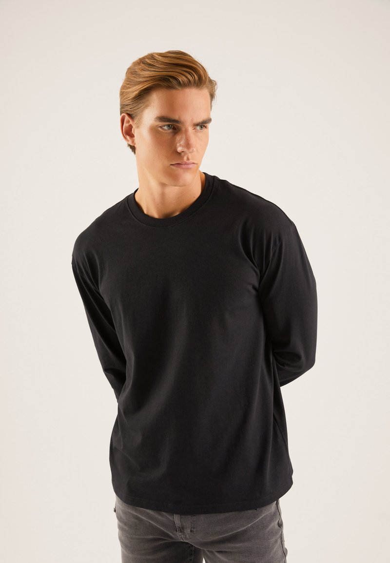 Tricou negru cu mânecă lungă din bumbac, având guler rotund și o croială lejeră. Asortat cu blugi gri, prezentat pe un fundal neutru.