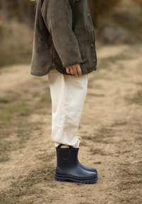 Marine rubberen regenlaarzen met gestructureerde oppervlakken, versterkte zolen en trekhandvatten, gecombineerd met witte broeken en een bruine corduroy jas.