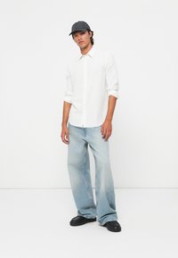 White button-up shirt with textured fabric, paired with light blue wide-leg jeans. Casual black cap and black shoes complete the look.