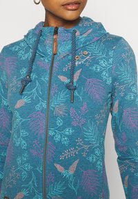 Mujer vestida con una chaqueta con cremallera azul y estampado floral en rosa, morado y azul claro, con cordones y pequeños botones de madera cerca del cuello.