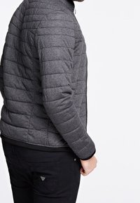 Grå quilted jakke med høj hals, polstret tekstur og lynlås lukning. Sorte detaljer ved ærmer og kant. Sorte bukser med sidelomme detalje.