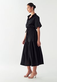 Juoda medvilninė suknelė su mygtukų apykakle, trumpomis rankovėmis, elastingu raišteliu liemeniui ir sluoksniuotu sijonu. Dėvima su nuogais šlepetėmis ant aukšto kulno.