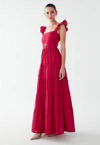 Vestido largo rosa de varias capas con mangas con volantes y escote cuadrado. Presenta un pequeño corte debajo del busto. Tela lisa con una textura suave.