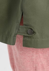 Manteau en tissu vert olive avec une fente latérale, doté d'une fermeture à pression argentée et d'une texture lisse. L'arrière-plan montre un textile rose.