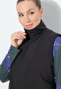 Schwarze ärmellose Weste mit hohem Kragen und vorderem Reißverschluss, kombiniert mit einem teal-farbenen Langarmshirt, das blaue und goldene abstrakte Muster aufweist.
