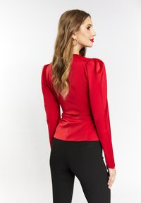Rød satinbluse med strukturerede skuldre og lange ærmer, tætsiddende ved taljen, parret med sorte leggings, set fra ryggen.