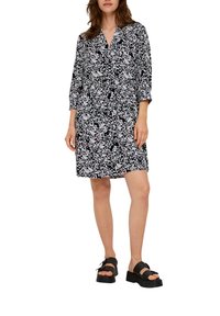 Schwarz-weißer, gemusterter Kleid mit V-Ausschnitt, Dreiviertelärmeln und lockerem Schnitt, kombiniert mit schwarzen Plateausandalen.
