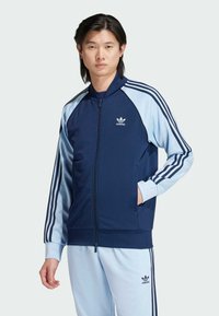 Námořnická modrá funkční bunda na zip s světle modrými rukávy a třemi bílými pruhy na každém rukávu; na hrudi je logo adidas.