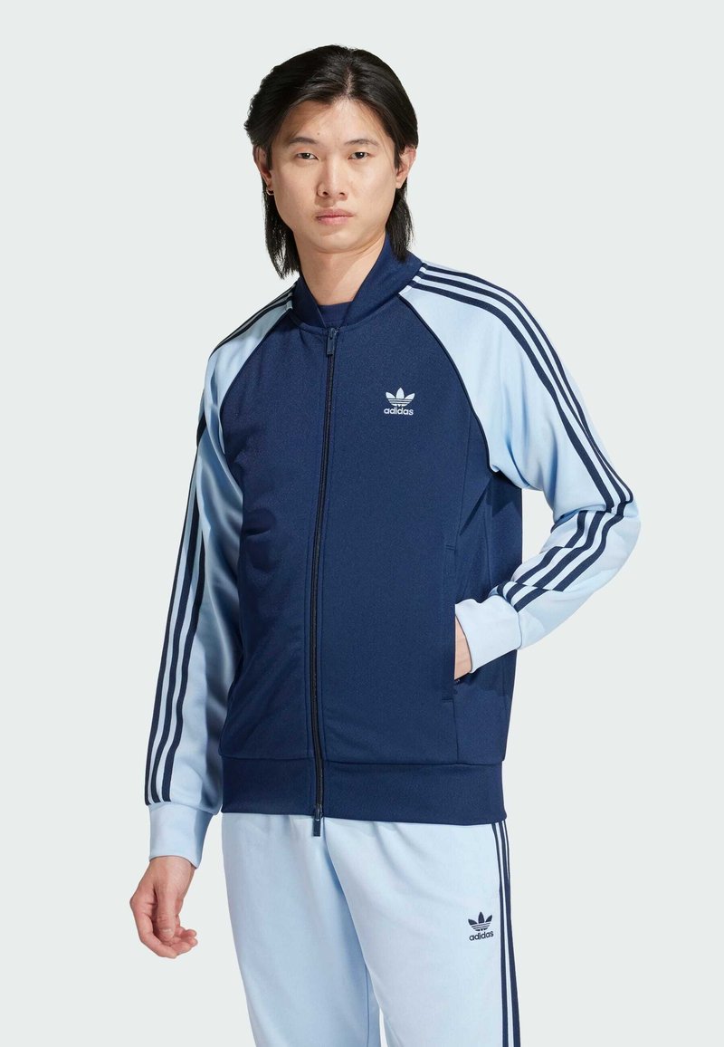 Námořnická modrá funkční bunda na zip s světle modrými rukávy a třemi bílými pruhy na každém rukávu; na hrudi je logo adidas.