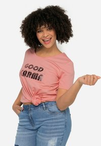 Camiseta rosa con "GOOD KARMA" en letras negras en negrita, anudada en la cintura. Combinada con jeans de mezclilla ligeros y desgastados. Cabello rizado.