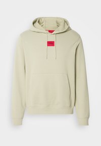 Sweat à capuche beige clair avec poche avant, cordons et petit patch logo rectangulaire rouge portant l'inscription HUGO sur la poitrine.