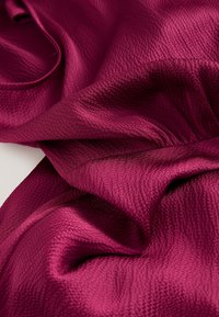 Fuksia tekstureret stof med et subtilt mønster, der falder i bløde, bølgede folder. Materialet virker let og glat.