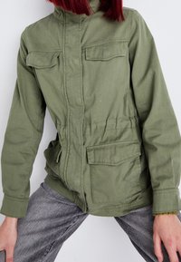 Oliivinvihreä puuvillatakki, jossa on korkea kaulus, etuvetoketju, kaksi rintataskua ja joustava vyötärö. Karkea kangas ja rento istuvuus.