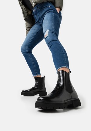 Personne portant un jean skinny bleu déchiré et des bottines Chelsea noires épaisses, debout avec une jambe en avant sur un fond blanc.