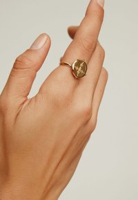 Anillo de oro con una piedra circular pulida que presenta elementos de diseño grabados, llevado en una mano mostrada sobre un fondo neutro.