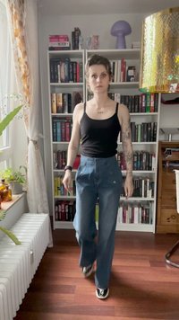Modre jeans hlače z visoko pasom in sprednjimi gumbi, v kombinaciji z črno majico brez rokavov. Notranji prostor z knjižnimi policami in rastlinami v ozadju.