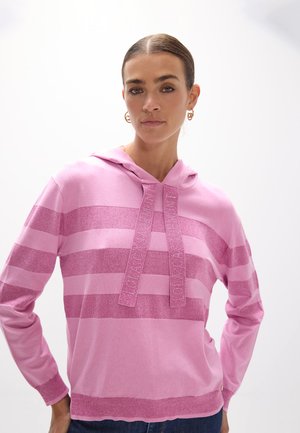 Mujer con el cabello peinado hacia atrás lleva una sudadera con capucha a rayas rosa con cordones y pendientes de aro dorados sobre un fondo blanco liso.