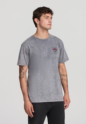 Uomo con capelli ricci e scuri che indossa una maglietta grigia sbiadita con una grafica di rosa rossa e jeans neri, in piedi su uno sfondo chiaro e uniforme.
