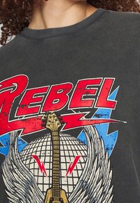 Harmaa puuvillapaita, jossa on graafinen suunnittelu, jossa lukee "REBEL" lihavoinnilla punaisella, sähkökitaran, siipien ja sinisten salaman aksenttien kanssa.