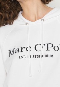 Sudadera blanca con cordón, que presenta un logo impreso en negro que dice "Marc O’Polo EST. IN STOCKHOLM" en el pecho.