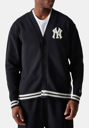 Mann trägt schwarzen geknöpften Cardigan mit weißem New York Yankees-Logo und gestreiften Bündchen, über einem weißen Hemd.