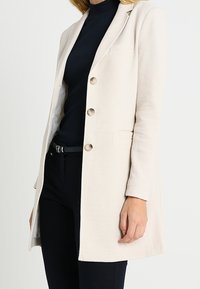 Beige dlouhý blazer s klopami, třemi knoflíky a postranními kapsami. Hladká tkanina a přizpůsobený střih. Nosený přes černý top.