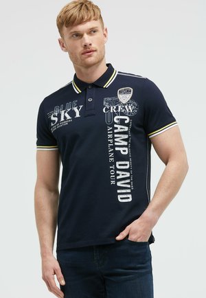 Junger Mann trägt ein marineblaues Poloshirt mit weiß-gelb gestreiftem Kragen und Ärmelbündchen, mit "Camp David" und luftfahrtthematischem Schriftzug.