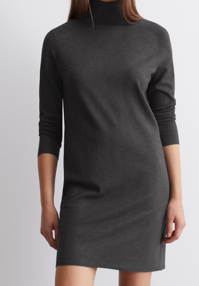Femme portant une robe-pull ajustée col roulé gris foncé à manches longues avec poignets côtelés, montrée des épaules jusqu’à mi-cuisse sur fond uni.