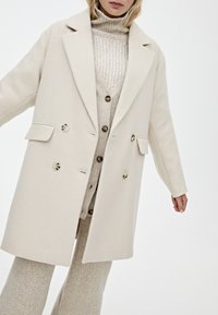 Cappotto beige doppiopetto con ampie rever, due tasche frontali e bottoni a contrasto. Indossato sopra un maglione lavorato a maglia beige chiaro e pantaloni.
