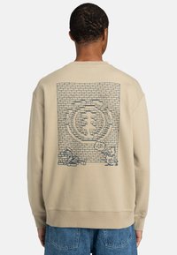 Sudadera beige con un diseño gráfico de una pared de ladrillos, personajes de dibujos animados y un globo de diálogo, tejido texturizado y puños acanalados.