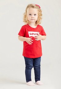 Red cotton t-shirt with a white logo, paired with dark blue skinny jeans. The child has curly hair and wears light socks.