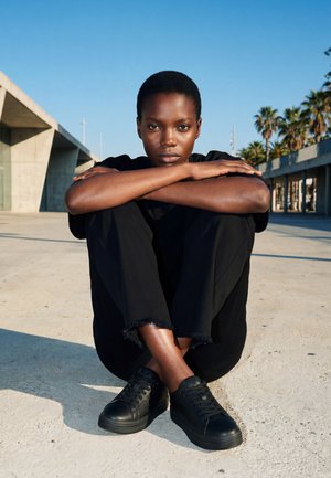 Młoda osoba o krótkich włosach w czarnych ubraniach i trampkach siedzi ze skrzyżowanymi nogami na betonie, z rękami opartymi na kolanach na zewnątrz.