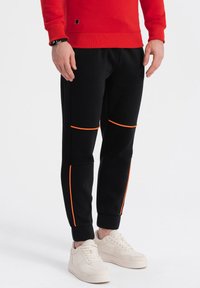 Sorte joggingbukser med orange accenter, strukturerede paneler og en tætsiddende pasform; båret med lyse sneakers og en rød sweatshirt.