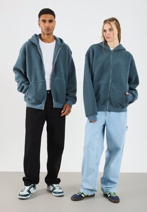 Deux personnes portent des vestes à capuche zippées surdimensionnées, fabriquées en tissu texturé et doux dans une couleur bleu atténué. Elles les assortissent avec un pantalon décontracté.