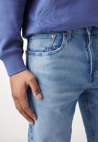 Person som bär ljusblå jeans och en lila sweatshirt med sin högra hand vilande mot sitt lår.